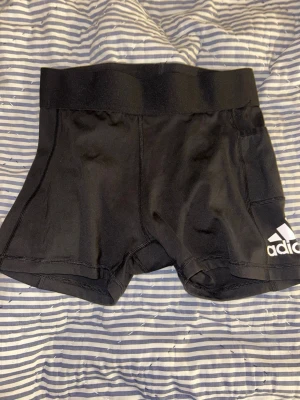 Svarta korta shorts Adidas - Säljer ett par svarta, tighta träningstights-shorts från Adidas med mellan hög midja och vit logga på benet. Gjorda i stretchigt funktionsmaterial (AEROREADY) som andas och sitter skönt på kroppen. Perfekta att ha under shorts eller bara som dom är .