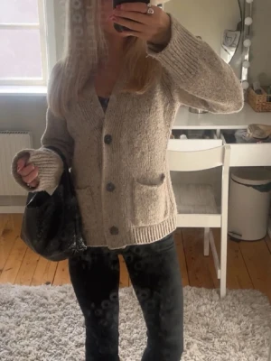 Beige kofta från Wera - Mysig beige stickad kofta med stora knappar framtill och två fickor. Koftan har en relaxed passform och ribbade muddar vid ärmslut och nederkant. Perfekt för lager på lager under kyliga dagar.