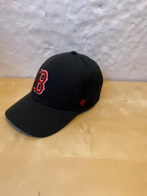 Svart Red Sox keps från '47 - Säljer en svart keps från '47 med klassiskt Red Sox B-broderi i rött och vitt framtill och 'RED SOX' broderat baktill. Justerbar passform och böjd skärm. Perfekt för dig som vill ha en sportig och stilren look.