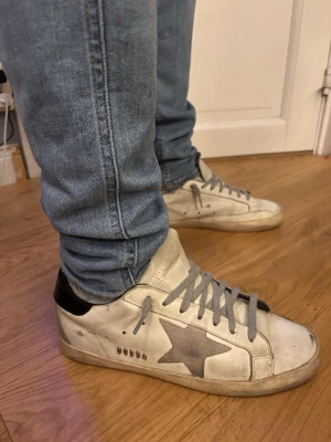 Golden goose - Golden Goose Superstar sneakers i vitt skinn med grå stjärna på sidan, grå snörning och svart hälparti. Klassisk låg modell med rund tå och platt sula. Snyggt slitna detaljer och retrovibe. Perfekta för dig som gillar streetstyle och vill sticka ut. Storlek 40 man passar 40-41