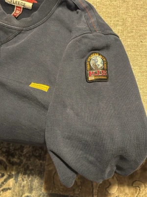 Mörkblå fleece tröja Parajumpers M - Mörkblå fleece tröja från Parajumpers i storlek M. Tröjan har en patch på ärmen och coola orange sömmar som ger extra detaljer. Klassisk rund halsringning och lång ärm, perfekt för dig som gillar stilrena och funktionella plagg med hög kvalité. Köpt för 2250kr