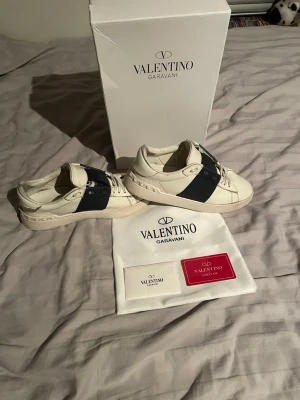 Valentino Garavani vita sneakers - Säljer ett par vita Valentino Garavani sneakers med bred mörkblå rem över sidan och ikoniska nitar runt hälen. Skorna har snörning och är tillverkade i skinn. Medföljer originalkartong, dustbag och äkthetskort. Perfekta för dig som gillar exklusiva detaljer och stilren design. Det är lite slitningar på dem 