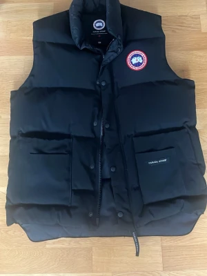Svart dunväst från Canada Goose M - Säljer en svart dunväst från Canada Goose i storlek M. Västen har två stora fickor framtill, hög krage och klassisk Canada Goose-logga på bröstet. Stängs med både dragkedja och tryckknappar. Perfekt för lager-på-lager under kyliga dagar.