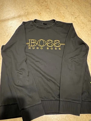 Svart Hugo Boss sweatshirt slim fit - Säljer en svart sweatshirt från Hugo Boss med guldfärgad logga framtill. Tröjan har rund hals, ribbade muddar och är i slim fit-modell. Perfekt för dig som gillar stilrena och klassiska plagg med en exklusiv touch.
