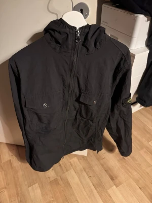 Svart vindjacka från Stone Island - Säljer en svart vindjacka från Stone Island med huva och dragkedja framtill. Jackan har två stora bröstfickor med knappar och den klassiska Stone Island-loggan på ärmen. Perfekt för dig som vill ha en clean och stilren look. Sälja billigt då jag behöver bli av med den. Kan mötas upp i Stockholm.