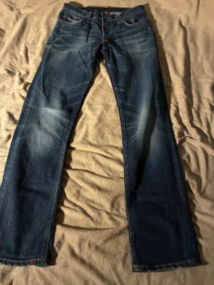 Blå jeans från Nudie Jeans, W30 L32 - Säljer ett par klassiska blå jeans från Nudie Jeans med raka ben och snygga slitningar. Jeansen har kontrastsömmar, stora bakfickor med broderad detalj och patch i läder bak. Perfekt passform och tidlös stil.