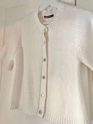 Vit stickad kofta från Gina Tricot - Säljer en söt vit stickad kofta från Gina Tricot med korta ärmar och rund hals. Koftan har ribbade kanter och knäpps med silvriga knappar framtill. Perfekt att slänga över en topp för en clean och fräsch look.