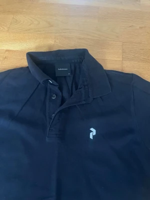 Svart pikétröja Peak Performance M - Klassisk svart pikétröja från Peak Performance i storlek M. Tröjan har korta ärmar, krage med knappar och en diskret logga på bröstet. Tillverkad i mjuk bomull som känns skön mot huden. Perfekt till jeans eller shorts för en clean look.
