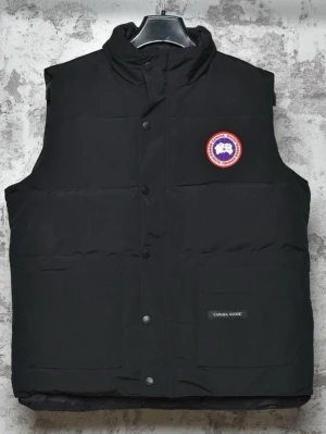 Svart dunväst från Canada Goose - Svart dunväst från Canada Goose med hög krage och tryckknappar framtill. Västen har två stora fickor och klassisk logotyp på bröstet. Perfekt för lager-på-lager och riktigt snygg till streetwear-stilen.