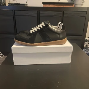 Maison Margiela sneakers svart - Maison Margiela sneakers i svart skinn och mocka med vita snören och vit insida. Klassisk gummisula i beige och diskret branding på sulan. Snygg retrostil med låg profil och rund tå, perfekta till streetwear eller casual outfits.