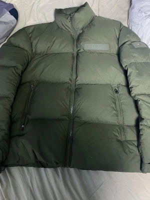 Grön pufferjacka från Tommy Hilfiger - Säljer en grön pufferjacka från Tommy Hilfiger med hög krage och två dragkedjeförsedda fickor. Jackan har diskret Hilfiger-logga på bröstet och är tillverkad i polyester med dunfyllning. Perfekt för kalla dagar och har en clean, modern look.