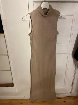 Beige ribbad långklänning NA-KD - Säljer en beige ribbad långklänning från NA-KD. Klänningen är ärmlös med hög krage och har en tight passform som framhäver figuren. Perfekt för dig som gillar stilrena och enkla plagg med en modern touch.