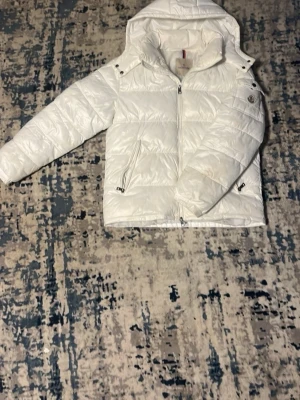 Vit Moncler pufferjacka med huva - Säljer en clean vit pufferjacka från Moncler med huva och tvåvägsdragkedja. Jackan har Moncler-logga på ärmen, praktiska fickor med dragkedja och quiltad design. Perfekt för kalla dagar och riktigt snygg till streetwear-looken.