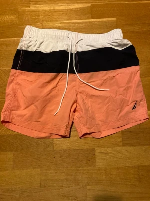 Randiga badshorts från Nautica M - Säljer ett par snygga badshorts från Nautica i storlek M. Shortsen har breda ränder i vitt, marinblått och korallrosa, med dragsko i midjan. Tillverkade i nylon med polyesterfoder, perfekta för stranden eller poolen.