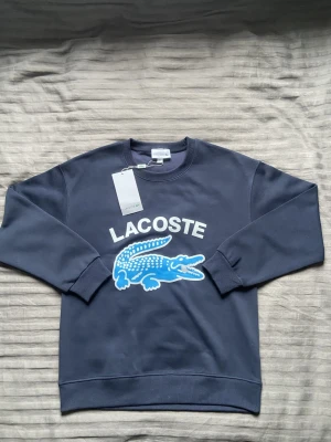 Mörkblå sweatshirt från Lacoste - PRIS KAN DISKUTERAS.Mörkblå sweatshirt med rund hals från Lacoste. Stor logga med vit text och blå krokodil framtill. Tröjan har ribbade muddar vid ärmslut och nederkant. Perfekt för en avslappnad och sportig stil.