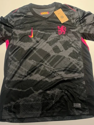 Chelsea matchtröja - Svart träningströja från Nike med Chelsea-logga och rosa detaljer. Mönstrad design i olika grå nyanser, rosa och gula inslag på sidor och loggor. Tillverkad i funktionsmaterial som andas, perfekt för sport och träning.