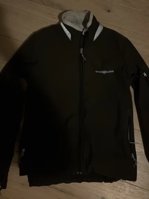 Svart skepparjacka från Henri Lloyd - Snygg svart skepparjacka från Henri Lloyd med dragkedja framtill, två sidofickor med dragkedja och vit logga på bröstet. Insidan har ett vitt meshfoder med svarta prickar och kragen har kontrasterande detaljer. Perfekt för dig som gillar stilren och sportig look.