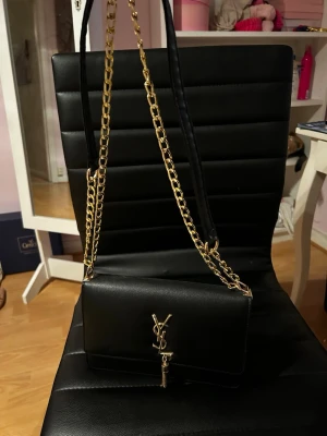 Svart YSL kuvertväska med guldkedja - Snygg svart kuvertväska från Yves Saint Laurent med guldfärgad kedjerem och ikonisk YSL-logga framtill. Väskan är i slätt läder och har en elegant tofsdetalj under loggan. Perfekt accessoar för att lyfta vilken outfit som helst. Helt oanvänd, pris kan diskuteras 🤍🤍🤍