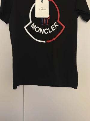 Svart Moncler t-shirt med logga - Svart t-shirt från Moncler med stor vit och röd logotyp på bröstet. Klassisk passform med korta ärmar och rund halsringning. Tillverkad i mjuk bomull som känns skön mot huden. Perfekt för dig som gillar stilrena och trendiga plagg.