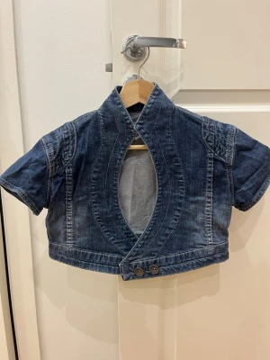 Kort jeansjacka med öppen front - Unik kort jeansjacka i blå denim med öppen oval front och två dekorativa knappar nedtill. Jackan har korta ärmar och markerad krage, perfekt för att ge outfiten edge. Snygga sömdetaljer framtill och klassisk jeanslook.