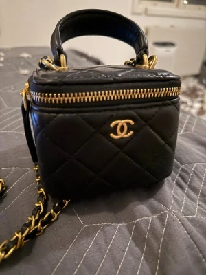 Chanel väska - Supersnygg svart quiltad miniväska från Chanel i äkta läder med guldfärgade detaljer och kedjerem. Väskan har klassisk CC-logga framtill, dragkedja upptill och vinrött tygfoder med innerfack. Perfekt accessoar för dig som gillar lyxig och trendig stil.