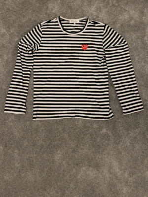 Randig långärmad tröja Comme des Garçons - Säljer en svartvit randig långärmad tröja från Comme des Garçons Play med det ikoniska röda hjärtat broderat på bröstet. Tröjan har rund halsringning och är gjord i mjuk bomull. Perfekt för dig som gillar streetstyle och vill ha något klassiskt men ändå lekfullt.