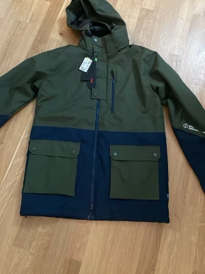 Grön och marinblå vindjacka Ski Industries - Stilren vindjacka från Ski Industries i mörkgrönt och marinblått. Jackan har huva, dragkedja framtill, två stora fickor med lock och tryckknappar samt en bröstficka med dragkedja. Perfekt för dig som vill ha en funktionell och snygg jacka till vardags.