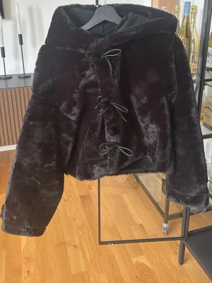 Pälsjacka  - Mysig svart fuskpälsjacka med stor luva med 3 knappar samt rosetter fram till. Endast använd 1 gång. 