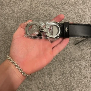 Svart bälte från Salvatore Ferragamo - Snyggt svart bälte från Salvatore Ferragamo i äkta skinn med en ikonisk silverfärgad dubbel-Gancini-spänne. Bältet har stilrena sömmar längs kanterna och klassisk design. Perfekt accessoar för att lyfta din outfit, ej plast mer, inte använd