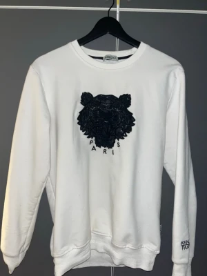 Vit Kenzo sweatshirt med tiger - Snygg vit sweatshirt från Kenzo med det ikoniska svarta tigerhuvudet broderat på bröstet och Kenzo Paris-text. Tröjan har rund hals, långa ärmar och ribbade muddar. Perfekt för dig som gillar streetwear och vill ha en statement piece.
