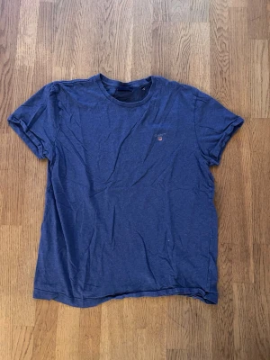 Blå t-shirt från GANT - En klassisk blå t-shirt från GANT med rund halsringning och liten broderad logga på bröstet. T-shirten har korta ärmar och är tillverkad i mjuk bomull, perfekt för en avslappnad och stilren look.