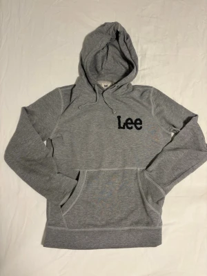 Lee Hoodie - Snygg grå hoodie från Lee med svart broderad logga på bröstet. Klassisk modell med huva och dragsnören samt stor magficka framtill. Tillverkad i mjukt sweatshirtmaterial, perfekt för en avslappnad stil.