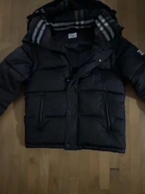 Burberry jacka  - Svart pufferjacka från Burberry med huva och klassiskt rutigt foder. Jackan har två stora fickor framtill, dragkedja och tryckknappar. På vänster ärm finns Burberry London England-tryck. Perfekt för kalla vinterdagar.