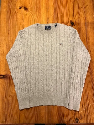 Ljusgrå kabelstickad tröja Gant - Kabelstickad tröja från Gant i ljusgrått med rund halsringning och diskret logga broderad på bröstet. Tröjan har långa ärmar och ribbade muddar vid ärmslut och nederkant. Perfekt för lager på lager och en clean look.