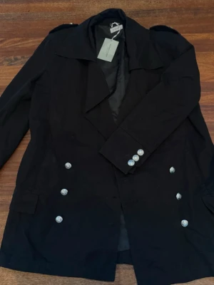 Svart dubbelknäppt rock från Hunkydory - Stilren svart rock från Hunkydory med bred krage och dubbelknäppning i silverfärgade knappar. Rocken har axelklaffar och två fickor framtill. Perfekt för dig som gillar klassisk men modern stil.