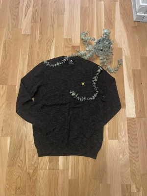 Mörkgrå stickad tröja Lyle & Scott - Mörkgrå stickad tröja från Lyle & Scott med rund halsringning och gul broderad logotyp på bröstet. Tröjan har en klassisk passform och ribbade muddar vid ärmslut och nederkant. Perfekt för lager-på-lager-stil.