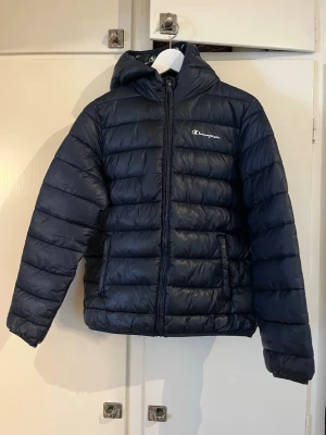 Mörkblå pufferjacka från Champion - Snygg mörkblå pufferjacka från Champion med huva och dragkedja framtill. Jackan har två sidofickor och diskret logga på bröstet. Quiltad design med breda kanaler och lätt glansigt material som ger en sportig vibe. Har en lite. Reva och vardags-slitningar annars i fint skick. Rök och Djurfritt. Barnstorlek! Stl. 13-14 år / 162/167 / ”X-Large” Mörk Marinblå