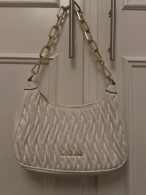  handväska Michael Kors - Snygg vit handväska från Michael Kors med quiltat mönster och guldfärgad kedja som axelrem. Väskan har en mjuk, rundad form och detaljer i guld, inklusive logotypen framtill. Perfekt accessoar för att lyfta din outfit.