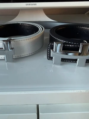 Svart och vitt bälte med H-spänne - Två stilrena bälten i svart och vitt med stora, blanka H-formade spännen i silver. Bältena har tydliga sömmar längs kanterna och är tillverkade i skinn. Perfekta för att lyfta din outfit med en modern touch.