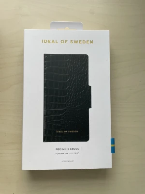 Svart croco mobilplånbok iDeal of Sweden - Stilrent svart mobilskal med croco-mönster från iDeal of Sweden. Passar iPhone 12/12 Pro och har plånboksfunktion. Tillverkat i konstläder med elegant struktur och guldfärgad logga. Perfekt för dig som vill ha både skydd och stil.