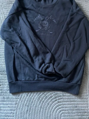 Sweatshirt från Adidas - Mörk marinblå sweatshirt från Adidas med broderad logga och två korsade flaggor på bröstet. Tröjan har rund halsringning och ribbade muddar vid ärmslut och nederkant.