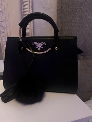 Svart handväska från Prada (ej riktigt!) - Snygg väska från Gran Canaria! svart, de övriga texten osv är guld färgat inte silver (de är ljuset). Den är precis som ny! Nån liten repa på metallhandtaget av att den legat i en garderob men inget som syns! Aldrig kommit till användning därför jag säljer, två snygga dekorationer och följer med ett axelband. Köpte för 450kr 