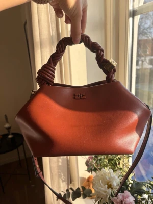 Brun handväska från Ganni i läder - Snygg brun handväska från Ganni i slätt läder med tvinnat handtag och justerbar axelrem. Väskan har guldfärgade detaljer och en stilren, modern form. Perfekt accessoar för att lyfta din outfit.