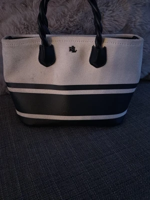 Randig handväska från Ralph Lauren - Snygg handväska i vitt och mörkblå från Ralph Lauren med tvinnade handtag och RL-loggan. breda ränder i mörkblått och vitt. Den har några fläckar men inget som jag tänker på och går säkert att tvätta bort. Orginalpris ca 1500 och är köpt i en riktig butik och är äkta. Priset går att diskutera 