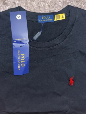 Svart t-shirt från Polo Ralph Lauren - Svart t-shirt från Polo Ralph Lauren med klassisk rund hals och röd broderad logga på bröstet. Tillverkad i mjuk bomull och har en normal passform. Perfekt för en stilren och enkel look.