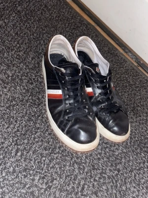 Moncler svarta lädersneakers - Snygga svarta sneakers från Moncler i läder med vita och röda detaljer på sidan samt klassisk logga på plösen. Skorna har svarta snören och vit sula som ger en clean look. Perfekta för dig som gillar stilrena och exklusiva sneakers.