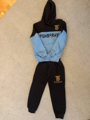 Burberry mjukisbyxor svart - Svarta mjukisbyxor från Burberry med guldig logga och text på vänster ben. Byxorna har resår i midjan med dragsko och muddar vid bensluten. Perfekta för en avslappnad och trendig stil. Passar BARN vs 6-8år size 6)