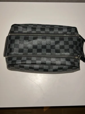 Svart necessär med rutigt mönster - Snygg necessär i svart och grått med klassiskt rutigt mönster och dubbla dragkedjor. Tillverkad i slitstarkt konstläder och har ett rymligt fack med svart tygfoder. Perfekt för att organisera dina småprylar eller smink. Praktiskt handtag på sidan för enkel transport.