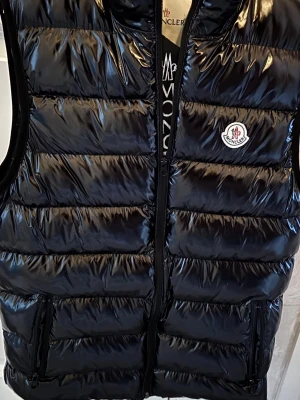 Svart dunväst  - Snygg svart dunväst med glansig finish och klassisk logga på bröstet. Västen har dragkedja framtill och två fickor med dragkedja. Perfekt för lager-på-lager och ger en cool streetwear vibe. Priset kan absolut diskuteras! 