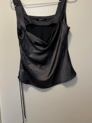 Svart satinlinnetopp från Gina Tricot - Snygg svart linnetopp i satin från Gina Tricot med draperad framsida och smala axelband. Toppen har en lätt glansig finish och snörning i sidan för en cool detalj. 